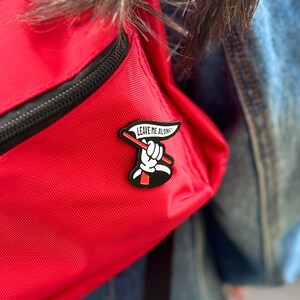 May include: A red backpack with a black zipper and a black and white enamel pin that says "Leave Me Alone!" with a hand holding a red flag.