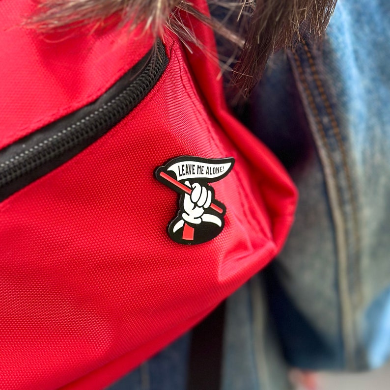 May include: A red backpack with a black zipper and a black and white enamel pin that says "Leave Me Alone!" with a hand holding a red flag.