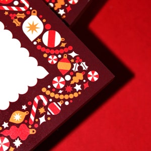 May include: Close-up of a dark red card with a white scalloped edge. The card is decorated with a festive border of red, white, and gold Christmas ornaments, candy canes, and stars. The background is a solid red.