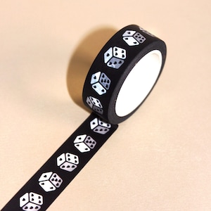 May include: A roll of black washi tape with a repeating pattern of white dice. The dice have gray shading to indicate the numbers. The tape is partially unrolled, revealing more of the dice pattern.