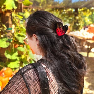 May include: A heart-shaped hair clip in black and red with a flame design. The hair accessory is worn in dark hair. The background shows a pumpkin patch with pumpkins and foliage.