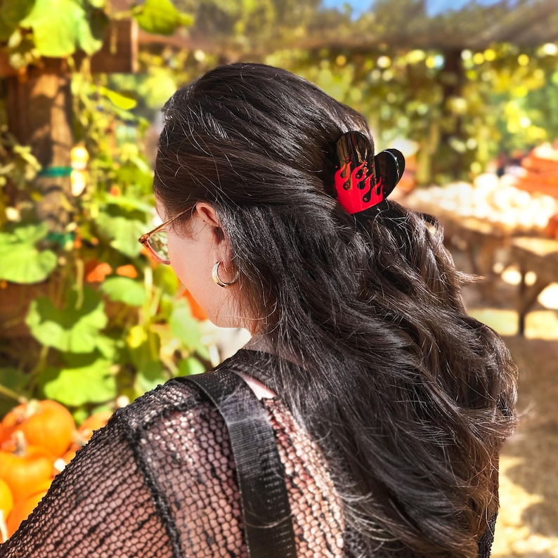 May include: A heart-shaped hair clip in black and red with a flame design. The hair accessory is worn in dark hair. The background shows a pumpkin patch with pumpkins and foliage.