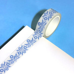 May include: A roll of decorative washi tape with a floral pattern in blue on a white background. The tape is partially unrolled, revealing the design. The background is a light blue.