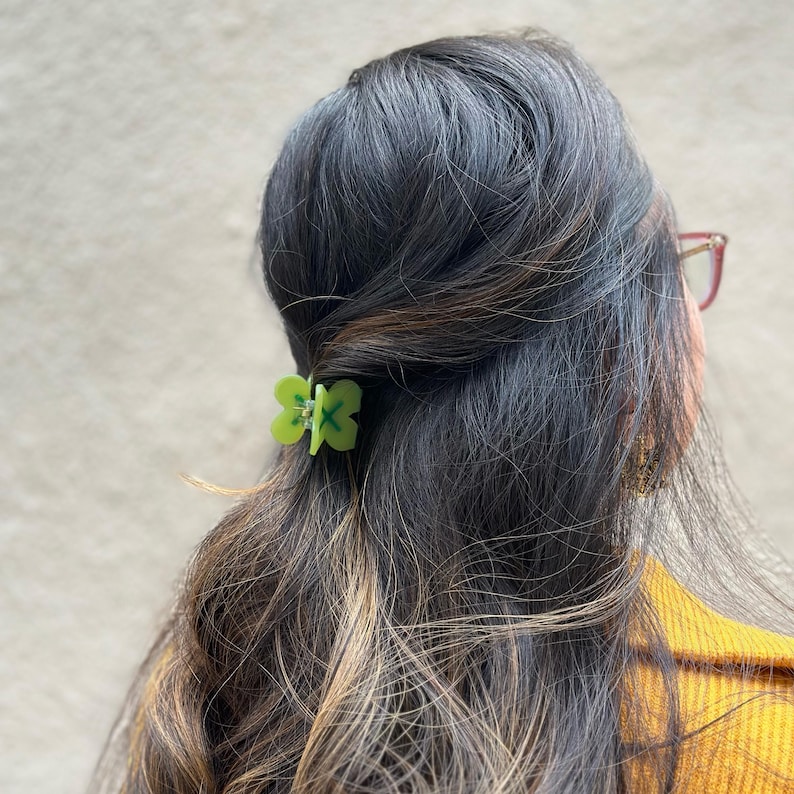 Pode incluir: Um grampo de cabelo verde de trevo de quatro folhas com um design em X verde e branco.