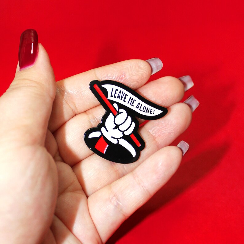 May include: A black and white enamel pin featuring a cartoon hand holding a white flag with the text "LEAVE ME ALONE!" in black lettering.