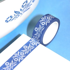 May include: A roll of blue washi tape with a white floral pattern. The tape is unrolled on a white surface, with a blue background. The floral design includes flowers and leaves, creating a decorative element for crafts and stationery.