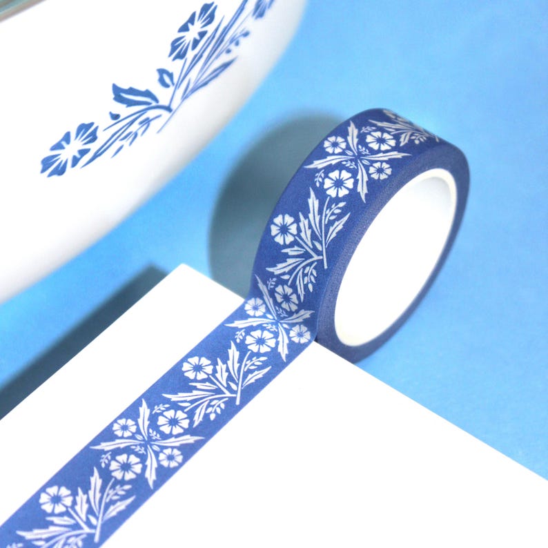 May include: A roll of blue washi tape with a white floral pattern. The tape is unrolled on a white surface, with a blue background. The floral design includes flowers and leaves, creating a decorative element for crafts and stationery.