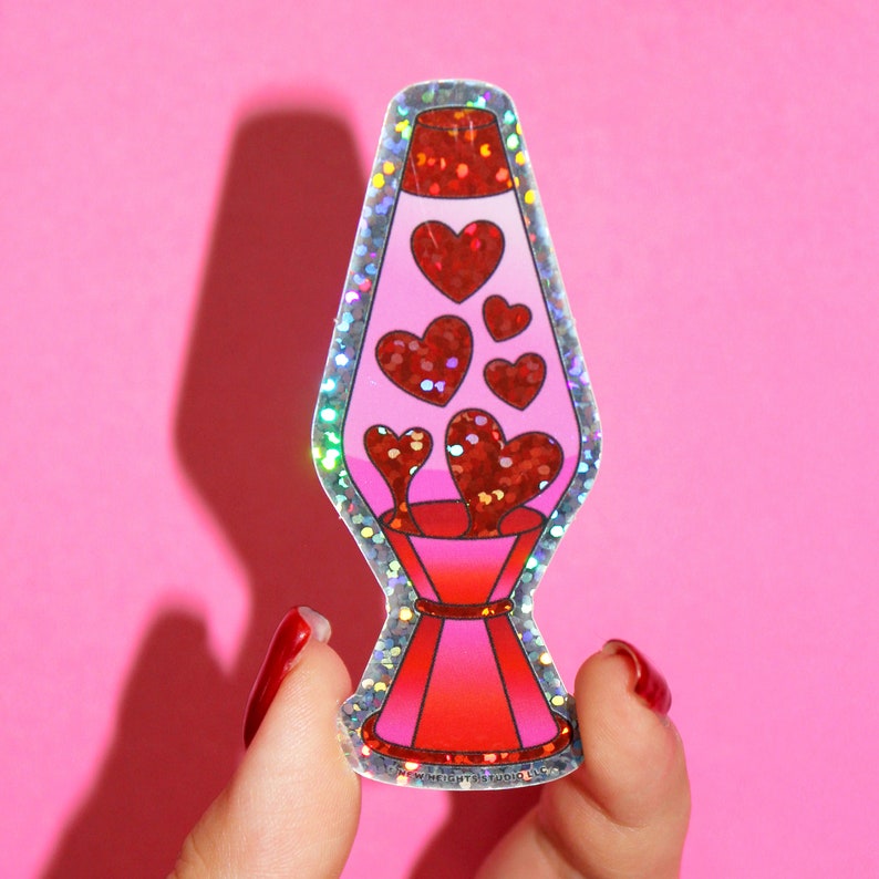 May include: A sticker featuring a lava lamp design with a pink body and red glitter hearts. The lamp has a red base and top, with a silver glitter border. The sticker is held against a pink background.
