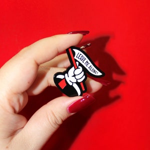 May include: A black and white enamel pin with a cartoon hand holding a red flag that says "Leave me alone!"