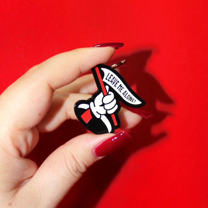 May include: A black and white enamel pin with a cartoon hand holding a red flag that says "Leave me alone!"
