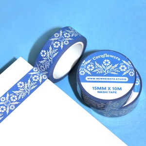 May include: Blue washi tape with a white floral pattern and the text "Cornflowers". The tape is 15mm x 10M. The tape is on a white surface against a blue background. The tape is from the New Heights Studio.