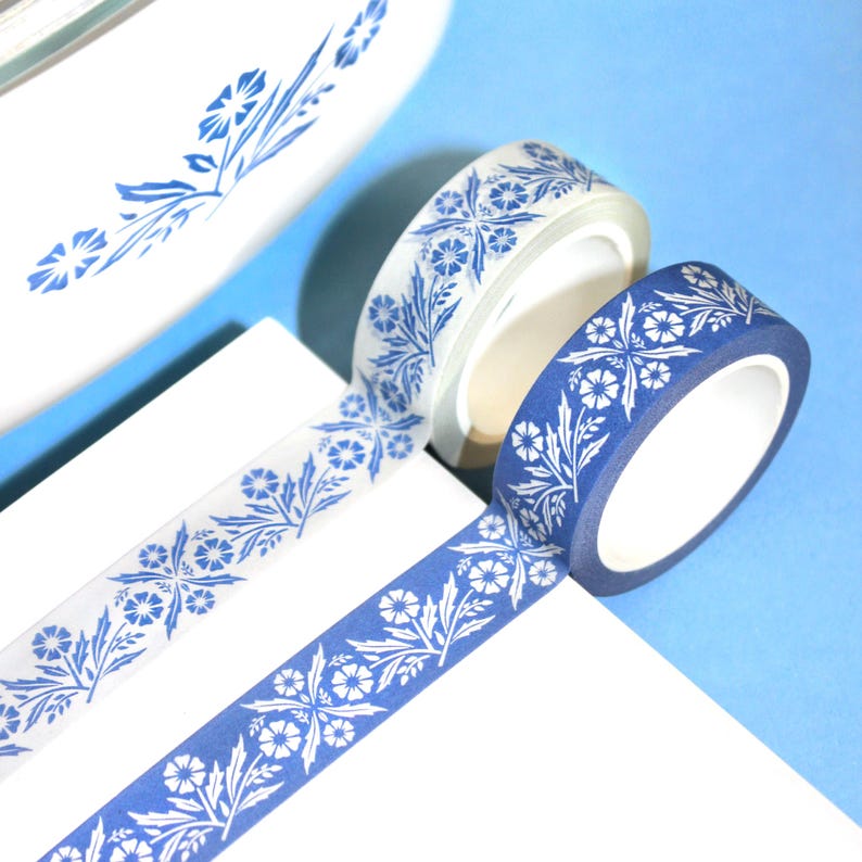 May include: Two rolls of decorative washi tape with a blue and white floral pattern. One roll is a deep blue, the other is a translucent white. The tape is unrolled on a white surface against a light blue background.
