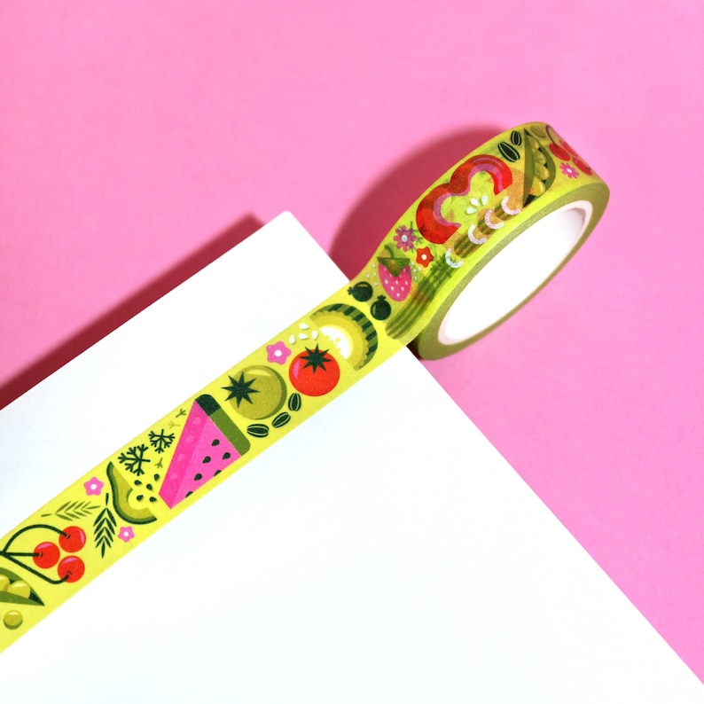 May include: A roll of yellow washi tape with a colorful pattern of fruits and vegetables. The tape is partially unrolled and is on a white surface.