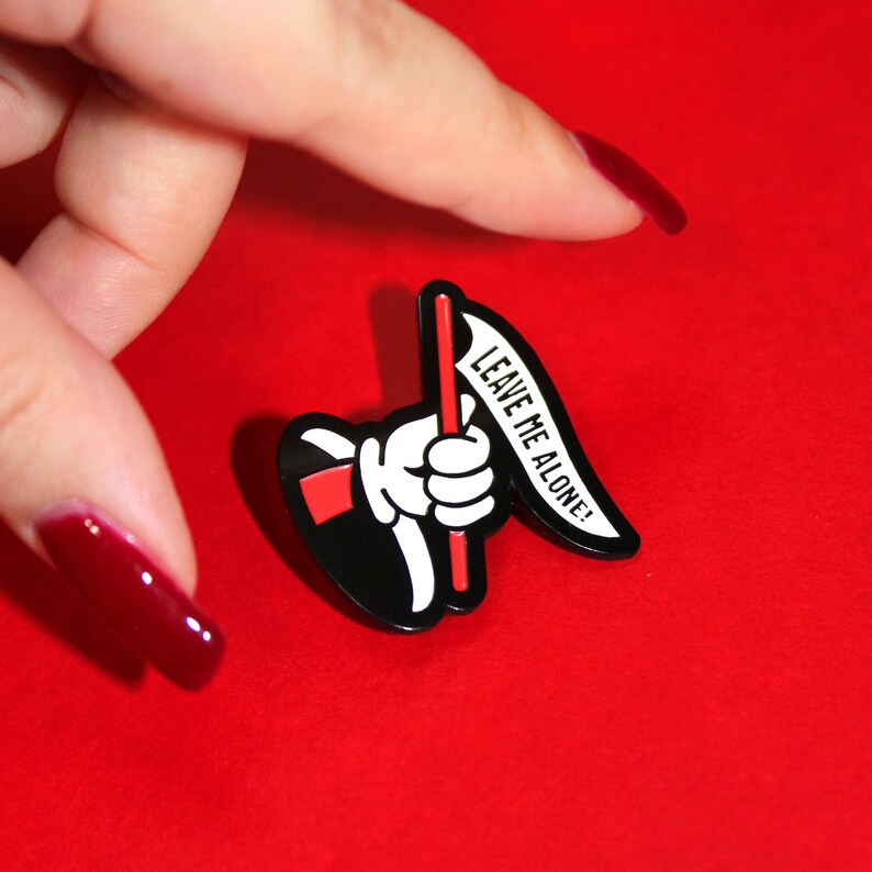 May include: Black and white enamel pin featuring a cartoon hand holding a red flag with the text "LEAVE ME ALONE!" in white.