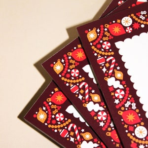 May include: A stack of Christmas cards with a dark red border featuring festive ornaments, candy canes, and starbursts in red, gold, and white. The cards have a scalloped edge and a blank white space for writing.