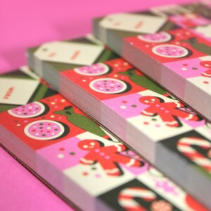 Peut inclure: Gros plan sur des piles d'&eacute;tiquettes cadeaux et de cartes de No&euml;l. Les cartes pr&eacute;sentent un motif r&eacute;p&eacute;t&eacute; de bonhommes en pain d'&eacute;pices, de cannes en sucre et de cercles roses et blancs sur un fond &agrave; carreaux rouges et blancs. Certaines &eacute;tiquettes portent le mot "FROM".