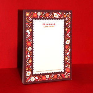 May include: A rectangular notepad with a festive Christmas border. The border features ornaments, candy canes, and stars in red, orange, and white against a dark brown background. The notepad has the text "FA LA LA LA LOTS TO DO" and is on a red surface.
