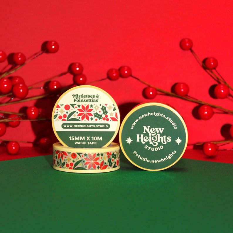 May include: Two rolls of washi tape with a Christmas theme. One roll features a green and red design with mistletoe and poinsettias. The other roll has a green and gold design with the text "New Heights Studio". Both rolls are 15mm wide and 10m long.