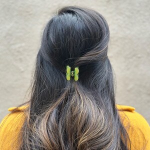 Pode incluir: Uma pessoa com cabelo comprido, castanho escuro com luzes, est&aacute; usando um grampo de cabelo verde. O grampo tem a forma de uma borboleta e tem um detalhe met&aacute;lico.