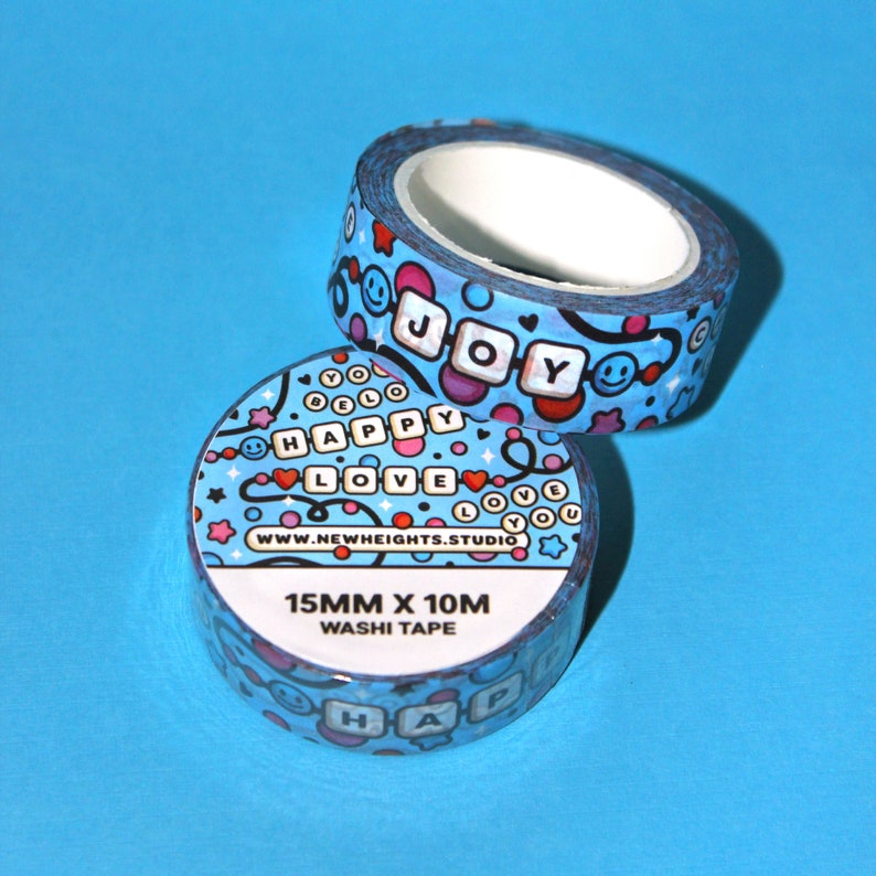May include: Two rolls of blue washi tape with a colorful pattern of hearts, stars, and smiley faces. The tape features the words "Happy", "Love", "Joy", and "You" in a blocky font. The tape is 15mm wide and 10m long. The website www.newheights.studio is printed on the tape.