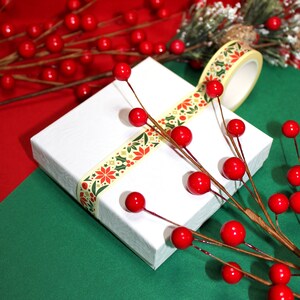 May include: A white gift box wrapped with a yellow and green patterned washi tape. The box is decorated with red berries and a sprig of greenery.