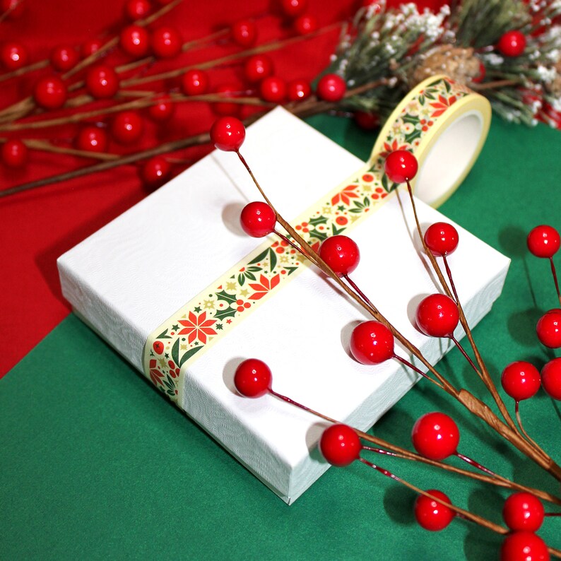 May include: A white gift box wrapped with a yellow and green patterned washi tape. The box is decorated with red berries and a sprig of greenery.