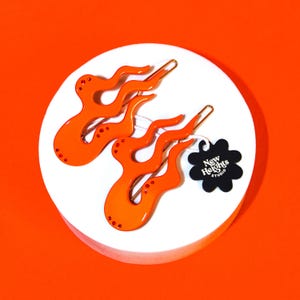 May include: Two orange flame-shaped hair clips with gold-colored metal pins, adorned with small, sparkling embellishments. The clips are displayed on a white circular surface against an orange background. A black tag with the text "New Heights Studio" is attached.