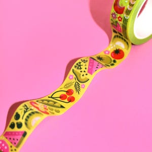 May include: A yellow washi tape with a colorful pattern of fruits, vegetables, and flowers. The tape is on a pink background.