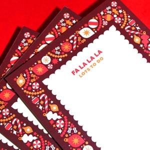 May include: A stack of burgundy notepads with a festive Christmas border. The border features ornaments, candy canes, and stars in red, orange, and white. The text "FA LA LA LA" and "LOTS TO DO" are printed in orange.