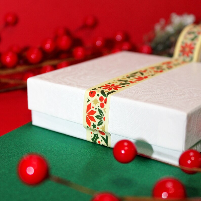 May include: A white gift box with a decorative ribbon featuring a red and green floral pattern. The box is sitting on a green surface with red ornaments.