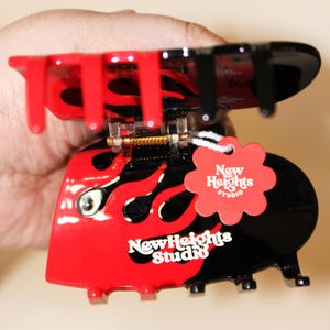 May include: A red and black hair claw clip with a flame design. The clip has the text "New Heights Studio" in white. A red tag with the text "New Heights Studio" is attached to the clip.