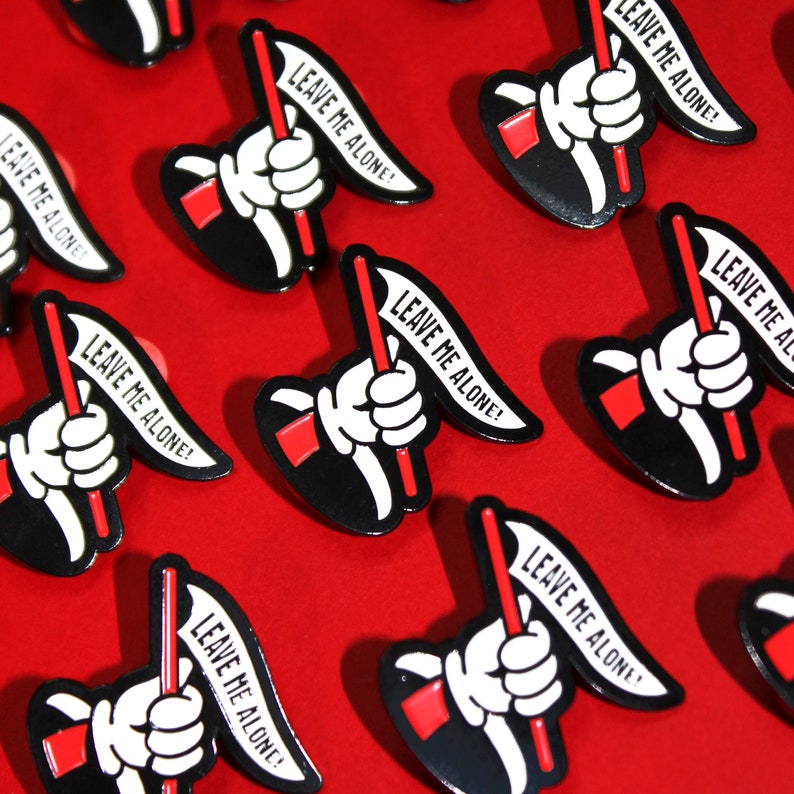 May include: A collection of black and white enamel pins with a red and white flag design. Each pin features a hand holding a flag with the text "Leave Me Alone!" on it.