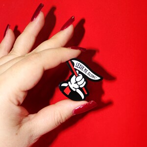 May include: Black enamel pin with a white hand holding a red flag that says "Leave Me Alone!"