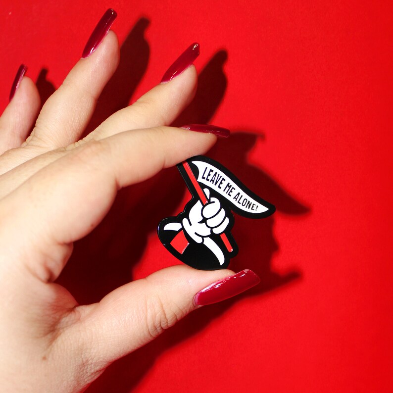 May include: Black enamel pin with a white hand holding a red flag that says "Leave Me Alone!"