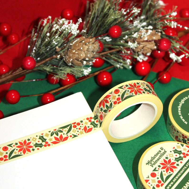 May include: Two rolls of washi tape with a Christmas theme. The tape features a repeating pattern of red poinsettias, green holly leaves, gold stars, and red berries on a cream background. The tape is rolled up and the ends are visible. The tape is on a green surface with red berries and a pine branch.