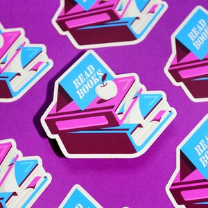 May include: A set of five stickers featuring a stack of books with a blue banner that says "Read Books" and a red apple on top. The stickers are on a purple background.