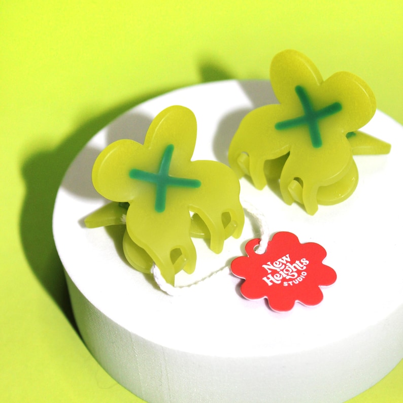 Pode incluir: Dois grampos de cabelo verdes em forma de trevo com um design de cruz verde. Os grampos est&atilde;o sobre uma superf&iacute;cie branca com uma etiqueta vermelha em forma de flor que diz "New Heights Studio".