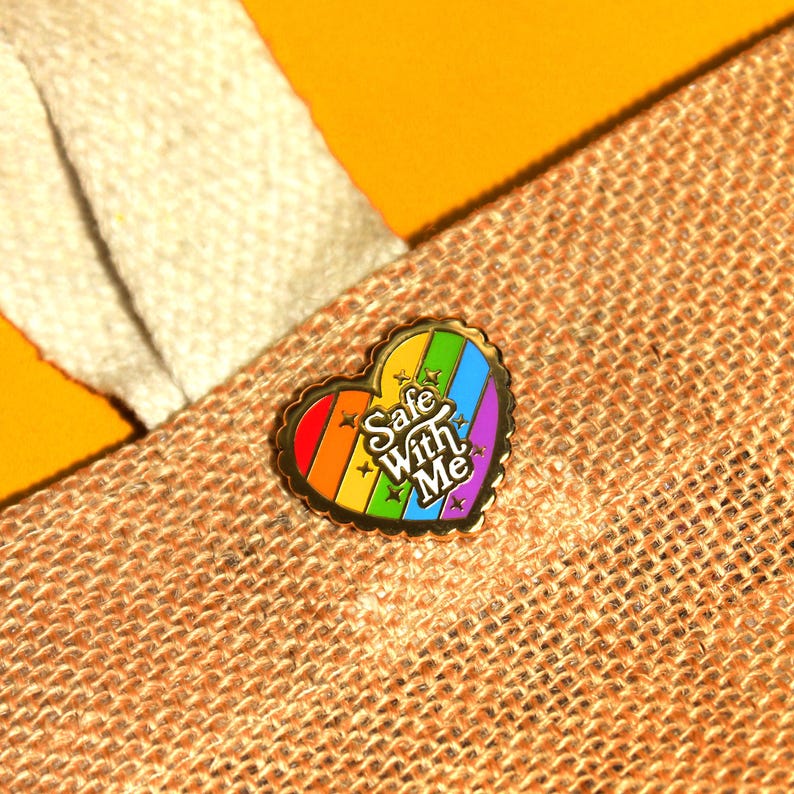 Op de afbeelding: Gouden hartvormige pin met een regenboogstrepenontwerp en de tekst "Safe With Me" in witte letters.