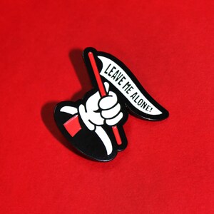 May include: Black and white enamel pin of a hand holding a flag that says "Leave Me Alone!" The flag is white with black text and the hand is white with black outlines. The pin is on a red background.