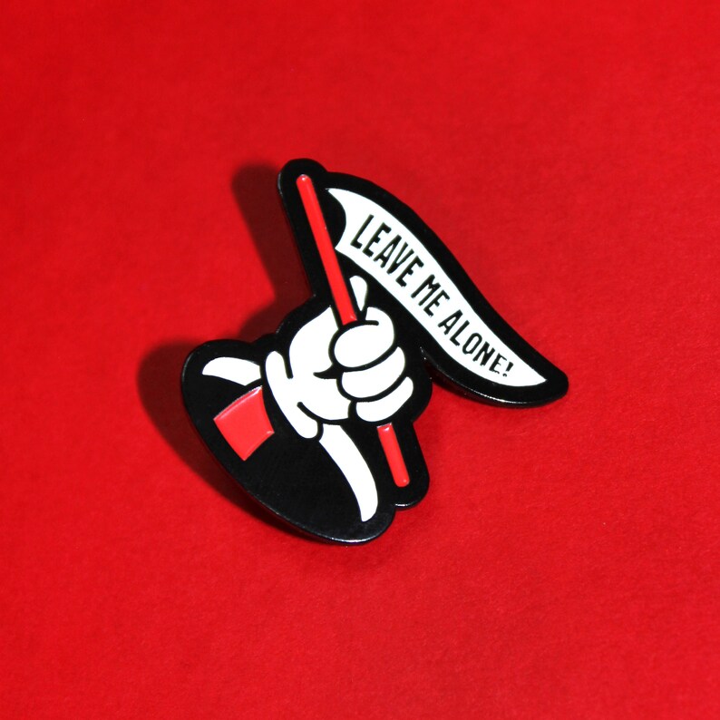 May include: Black and white enamel pin of a hand holding a flag that says "Leave Me Alone!" The flag is white with black text and the hand is white with black outlines. The pin is on a red background.