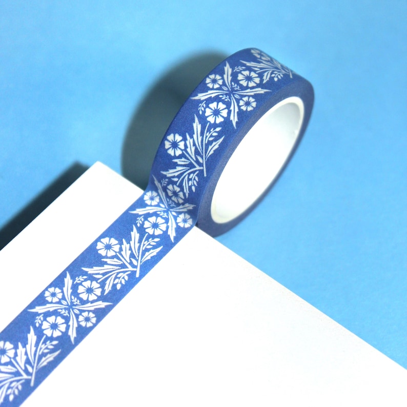 May include: A roll of blue washi tape with a white floral pattern. The tape is unrolled slightly, revealing the white interior of the roll. The background is a light blue.