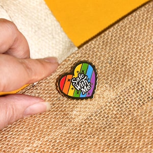 Op de afbeelding: Een gouden hartvormige emaille pin met een regenboogstreep ontwerp en de tekst "Safe With Me" in zwarte letters.
