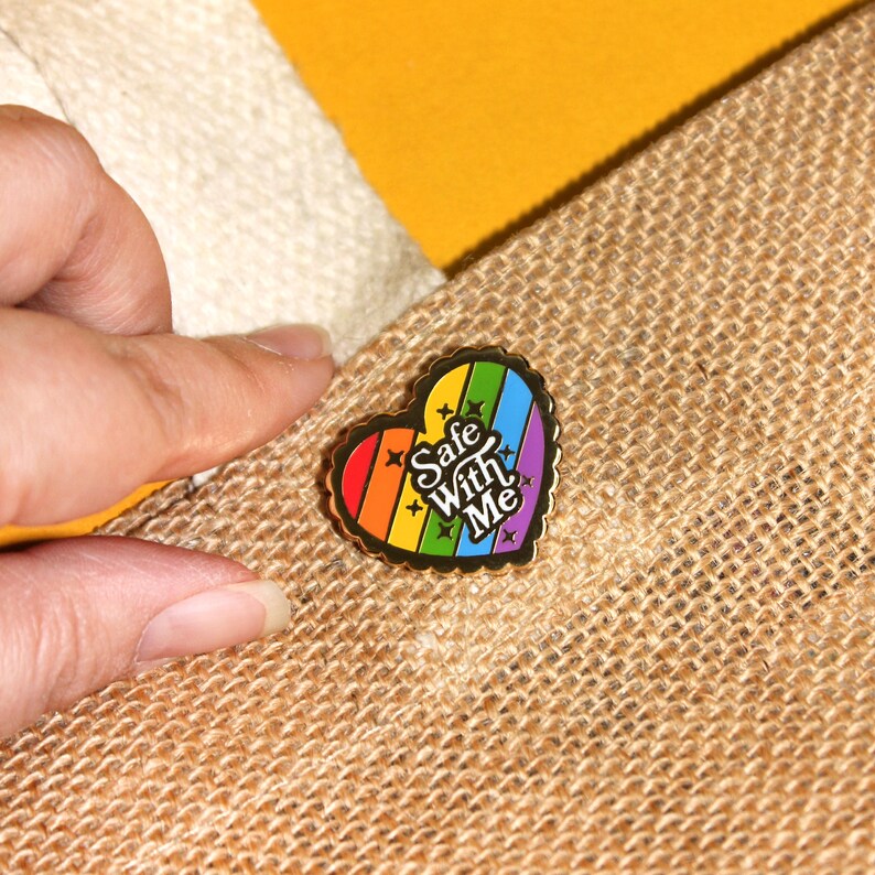 Op de afbeelding: Een gouden hartvormige emaille pin met een regenboogstreep ontwerp en de tekst "Safe With Me" in zwarte letters.