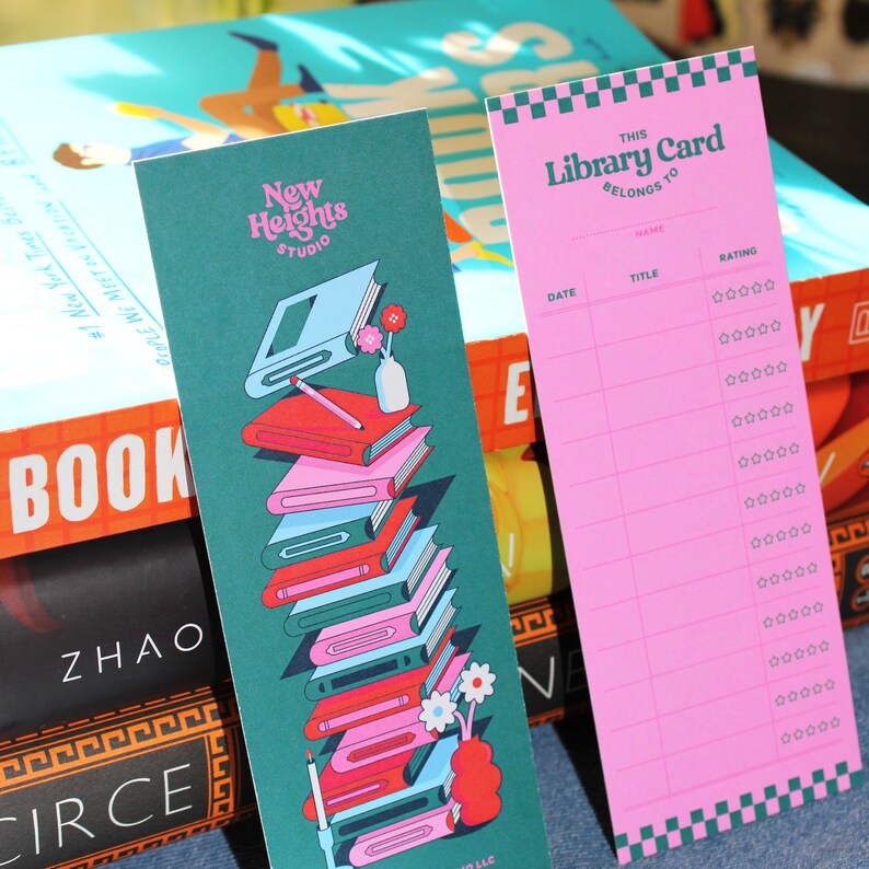 May include: A pink bookmark with a green and white checkered border. The bookmark has the text "This Library Card Belongs To" and a table with columns for "Date", "Title", and "Rating". The rating column has five stars for each row.