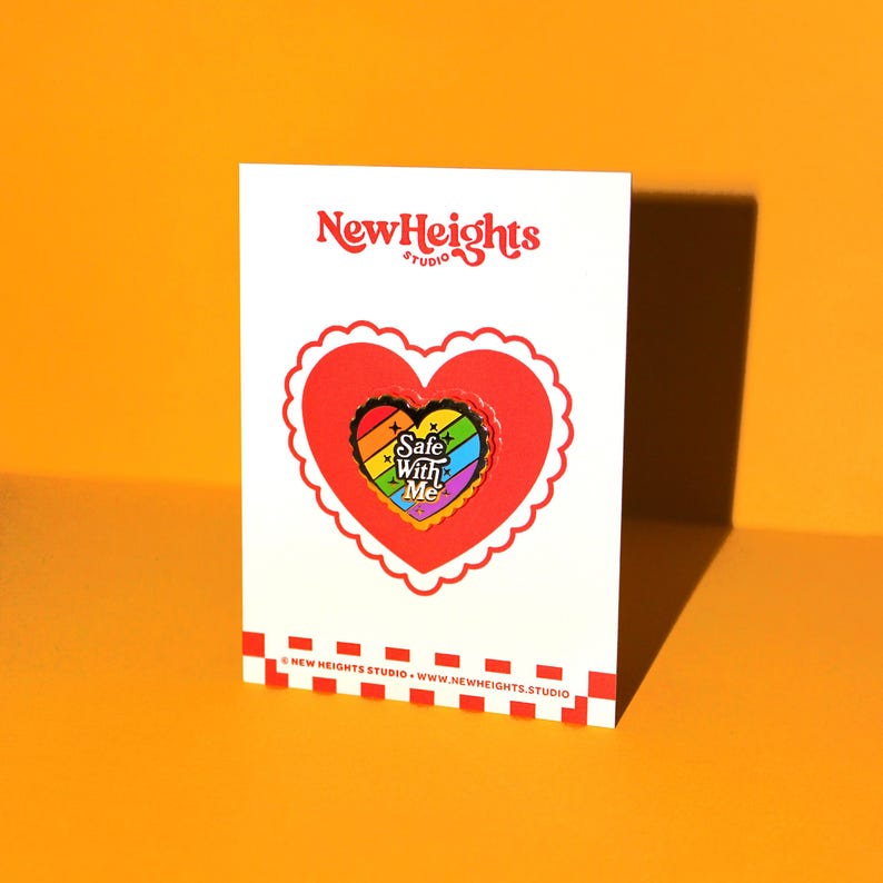 Op de afbeelding: Een regenboogkleurige hartvormige emaille pin met de tekst "Safe With Me" op een witte kaart met de tekst "NewHeights Studio" in een rood hart.