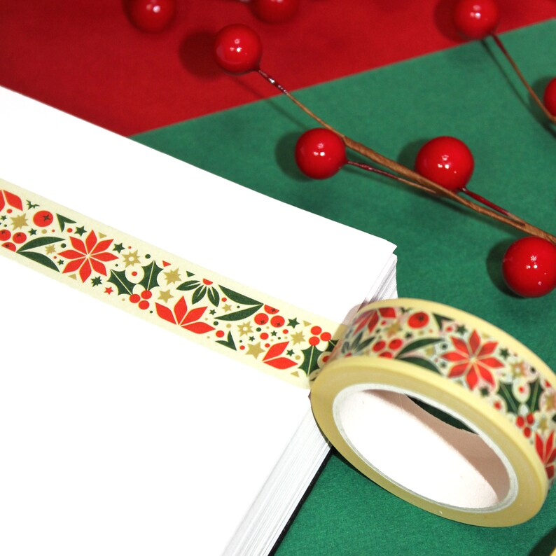 May include: A roll of washi tape with a Christmas-themed design featuring red poinsettias, green holly leaves, gold stars, and red berries on a cream background.