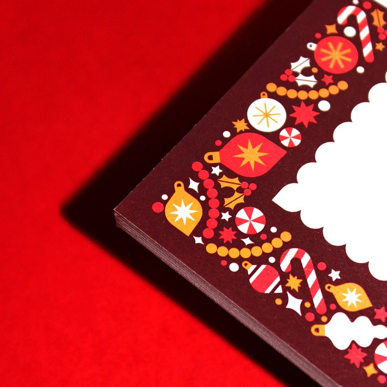 May include: A stack of Christmas cards with a festive border design. The border features ornaments, candy canes, stars, and holly in red, white, and gold on a dark brown background. The cards are on a red surface.