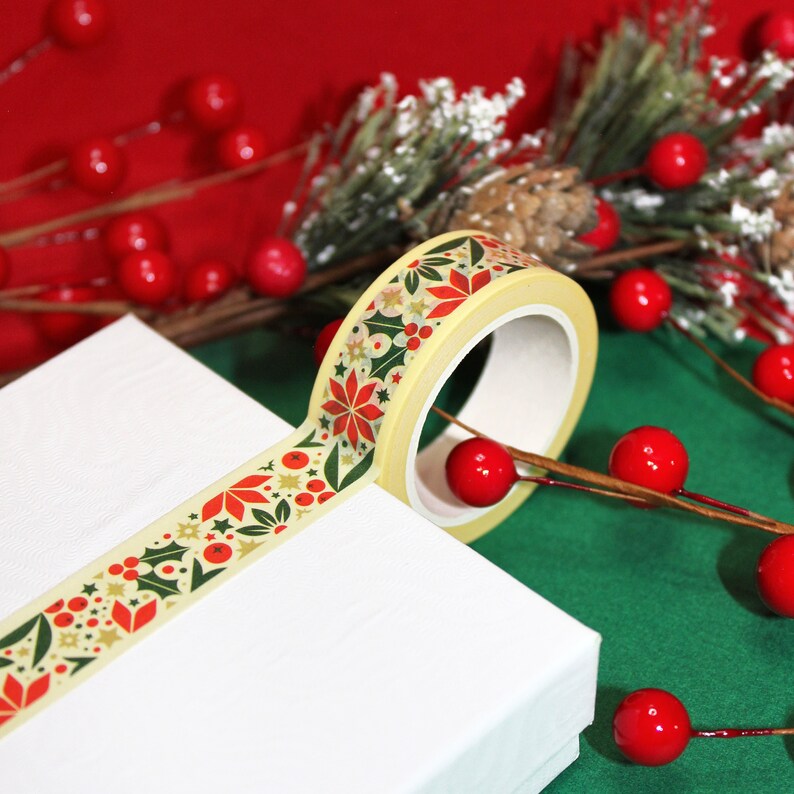 May include: A roll of washi tape with a Christmas-themed design featuring red poinsettias, green holly leaves, and gold stars on a cream background.