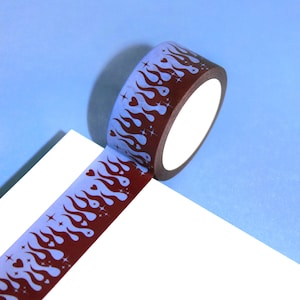May include: A roll of decorative washi tape featuring a repeating pattern of flames, hearts, and stars in shades of brown and light blue. The tape is angled on a white surface against a light blue background. Ideal for scrapbooking and crafts.