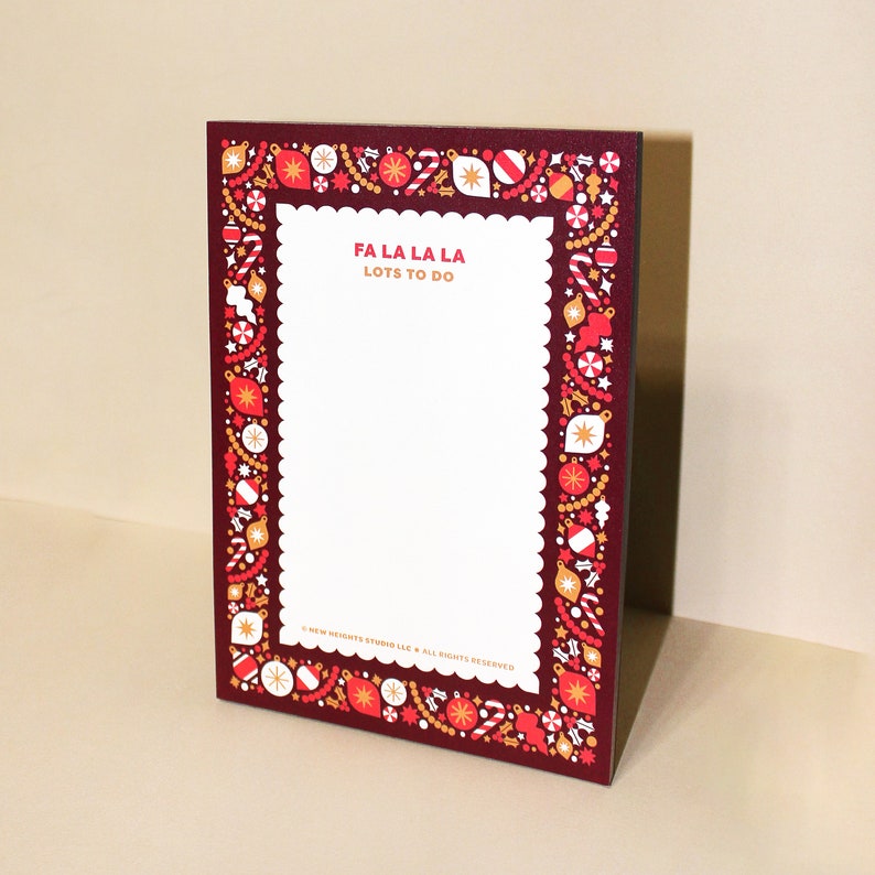 May include: A rectangular notepad with a burgundy border decorated with Christmas ornaments. The notepad has a white interior with the text "FA LA LA LA LOTS TO DO" in orange. The notepad is a festive holiday item.